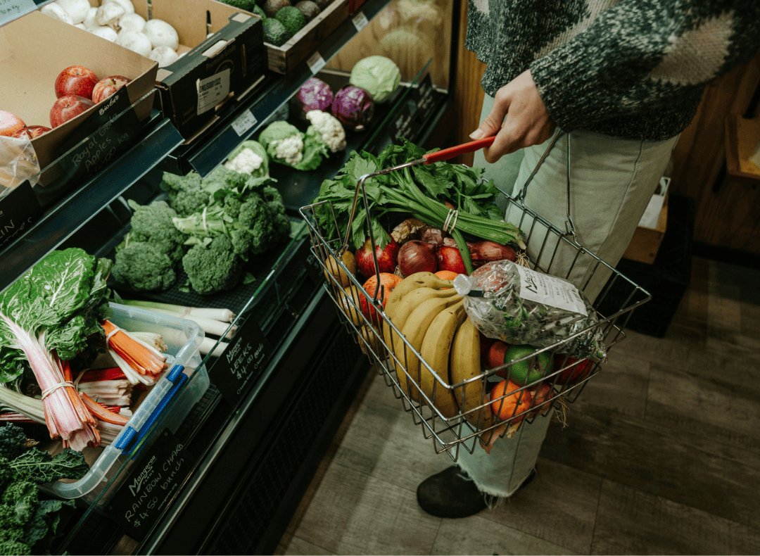 A shopper filling a wire basket with bananas, apples, celery and fresh greens at the Mangaroa Farm Shop produce shelves